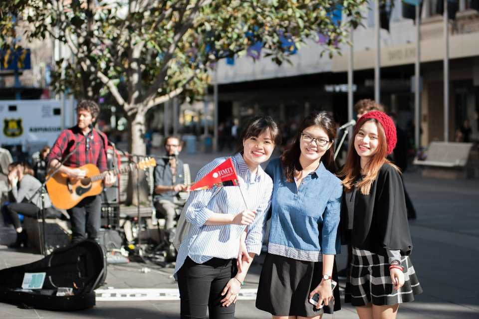 three female Vietnamese students waving RMIT flag on the streets of Australia in front of a busking band