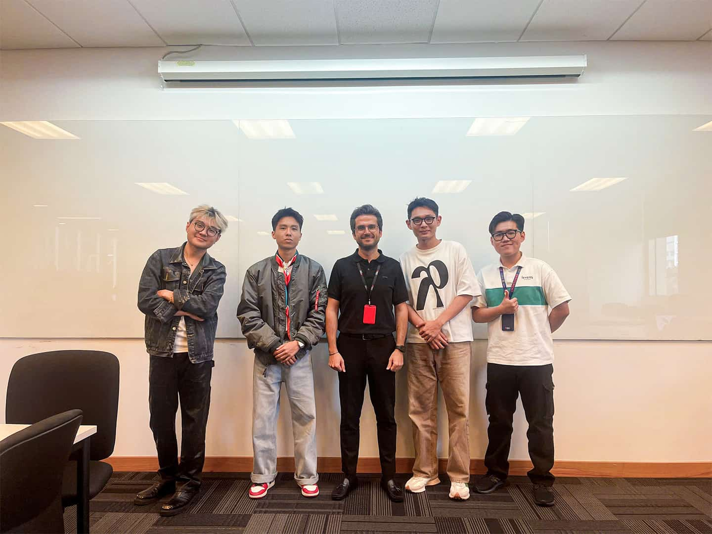 4 Vietnamese design students, including Nam Bao, winner of the design competition, wearing faded blue jeans, a grey jacket and red and white shoes with a vermillion cravat and a dress watch, standing with the design studies lecturer Abbas who is dressed in a black polo, black skinny jeans and black dress shoes. He is bearded and wears black wire-rimmed glasses, and a black Casio watch. 