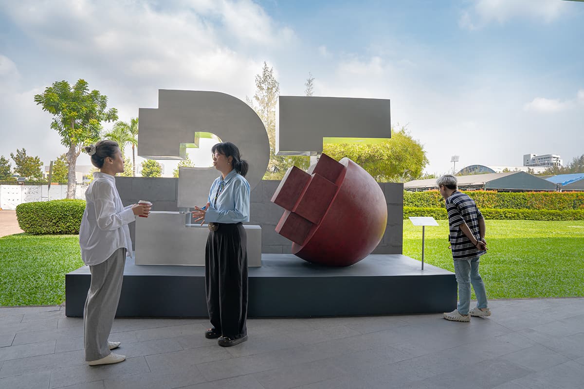 3 people chatting in front of the anniversary sculpture ourtdoors- sculpture is a three dimensional logo in the shape of a pixelated "25", with the round section of the "5" replaced with a three dimensional spherical imagination of the RMIT pixel. 