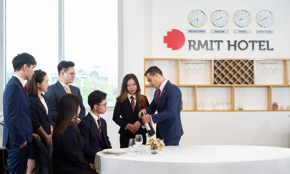 A group of students in business attire standing next to a professor demonstrating how to open a wine bottle in the mock RMIT Hotel